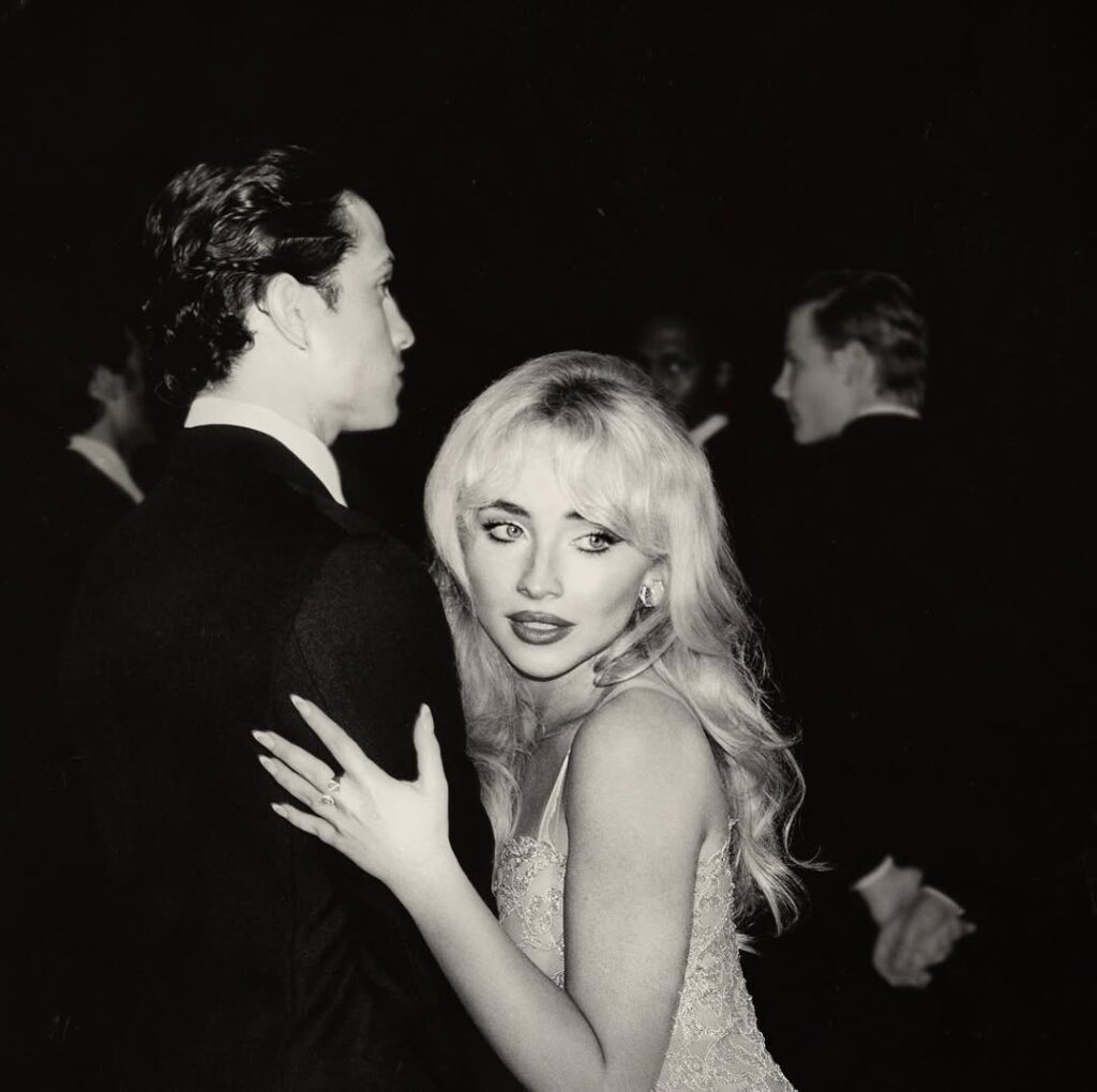 Black-and-white photo of Sabrina Carpenter in an embellished gown, embracing a man in a suit while glancing back toward the camera at a formal event.