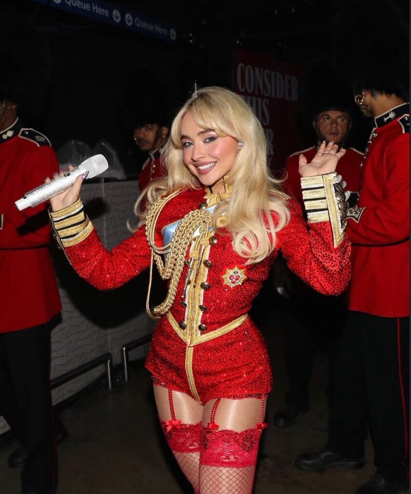 Sabrina Carpenter smiling backstage in a red sequined British guard–inspired outfit with gold embellishments and red lace stockings, holding a sparkly microphone.